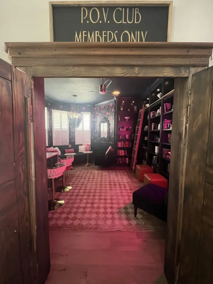 An antique armoire with its doors open beneath a 'P.O.V. Club, Members Only' sign, revealing a hidden home library glowing with pink light — the entrance to Pillars of Vices Book Bar